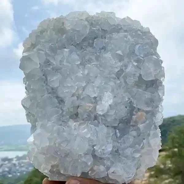 Large crystal cluster held against a scenic outdoor background with mountains and water.